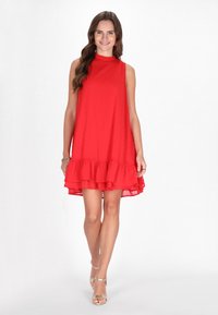 Rotes ärmelloses Kleid mit hohem Halsauschnitt, strukturiertem Stoff und einem Rüschensaum. Das Kleid endet über dem Knie. Kombiniert mit silbernen Sandalen mit Riemchen.