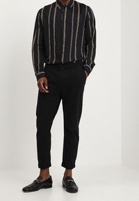 Zwarte overhemden met verticale witte en gouden strepen, gecombineerd met zwarte tapered trousers en glanzende zwarte loafers.