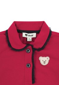 Red baby shirt with black trim, two black buttons, and a small teddy bear patch near the chest, size 62 for 2-3 months old.