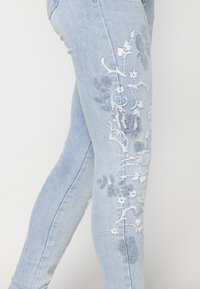 Jean skinny bleu clair avec broderies florales et papillons blanches et grises sur le côté de la jambe.