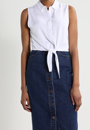 Biała, bezrękawnikowa koszula na guziki, wiązana w talii, zestawiona z wysokim stanem, ciemnoniebieską spódnicą dżinsową na guziki z przodu.