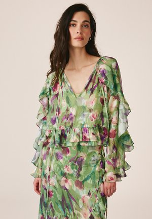 Femme aux longs cheveux foncés portant une robe verte transparente à motifs floraux avec des manches à volants et un décolleté en V sur un fond clair uni.