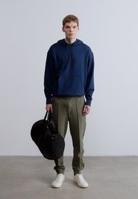 Tengerészkék kapucnis pulóver kenguruzsebbel, olívazöld nadrág és fehér tornacipő. A modell fekete sporttáskát tart a kezében. Sima anyagtextúra.