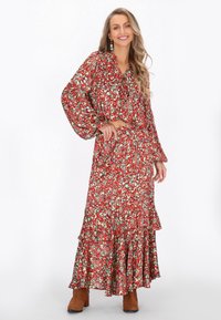 Lange rode bloemenjurk met groene accenten, gerimpelde zoom en losse mouwen. Model draagt bruine enkellaarsjes en hangende oorbellen.