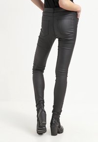 Jeans skinny de cuero sintético negro con una textura suave, cintura media, y bolsillos traseros, combinados con botas de tacón a la altura del tobillo.