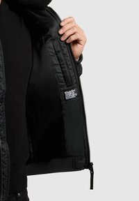 Giacca bomber nera con esterno trapuntato, chiusura con zip e interno morbido. Presenta un'etichetta con logo sul lato sinistro della fodera interna.