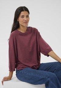 Donna con lunghi capelli scuri è seduta su un blocco bianco, indossa un maglione largo color bordeaux e jeans blu, contro uno sfondo chiaro uniforme.
