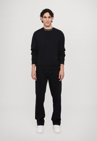 Felpa nera con polsini e scollatura a coste, caratterizzata da testo bianco. Abbinata a pantaloni neri e sneaker bianche, in piedi su uno sfondo neutro.