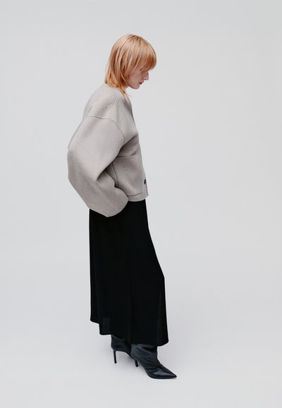 Hellgraue, oversized Wollejacke mit abgerundeten Schultern, kombiniert mit einem schwarzen Rock und spitzen schwarzen Stiefeln mit Absatz, vor einem neutralen Hintergrund.