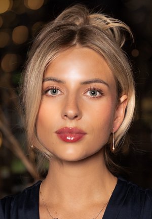 Eine Person mit blonden Haaren und einer eleganten Frisur, die ein dunkles Oberteil trägt, glänzenden rosa Lippenstift und tropfenförmige Ohrringe mit einem dezenten Glanz.