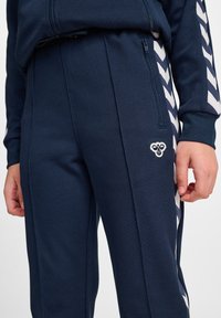  marineblauwe sportbroek van textuurstof, met witte herringbone zijstrepen en een klein logo op de linker bovenbeen.