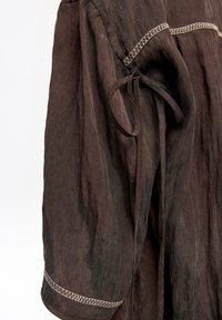 Brązowy, lekki materiałowy element garderoby o fakturowanej powierzchni, z zapięciem na sznurek oraz ozdobnym przeszyciem wzdłuż szwów i mankietów.