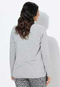 Donna con lunghi capelli ondulati scuri, che indossa una maglietta a maniche lunghe grigia chiara e pantaloni con stampa leopardata, in piedi contro una parete bianca di profilo.