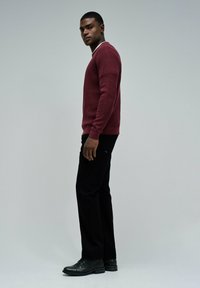 Jeune homme en pull bordeaux, pantalon noir et chaussures habillées noires se tenant de profil devant un fond gris uni.