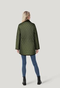 Olivgrüner gesteppter Mantel mit einem langen Design, ausgestattet mit einem gerippten Kragen und weiten Ärmeln, kombiniert mit blauen Slim-Fit-Jeans und schwarzen Stiefeln.