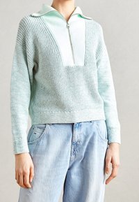 Minzgrüner Strickpullover mit halbhohem Reißverschlusskragen und strukturiertem Muster; getragen über hellblauen Jeans.