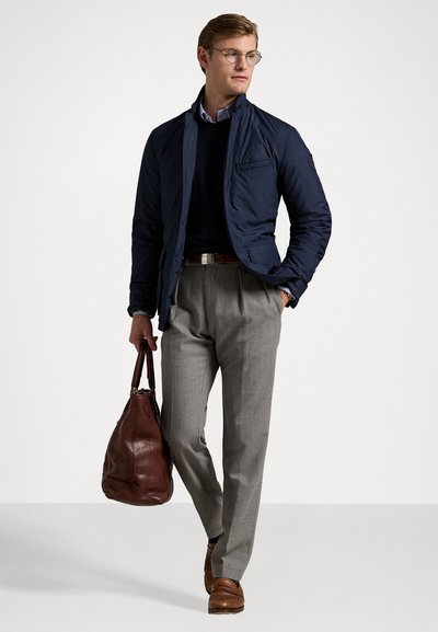 Chaqueta acolchada azul marino sobre un suéter oscuro, pantalones grises, zapatos de cuero marrón y un bolso de cuero marrón. Presenta gafas y una camisa a rayas.