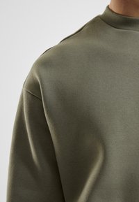 Olivenfarvet trøje med en glat tekstur, rund halsudskæring og subtile skuldersømme. Stoffet fremstår let og strækbart.