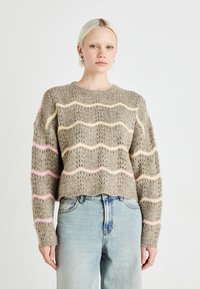 Gebreide trui in lichtbruin met golvende pastelstrepen (roze, geel) en opengewerkte patronen. Ronde halslijn en een cropped zoom. Gecombineerd met lichtblauwe jeans.