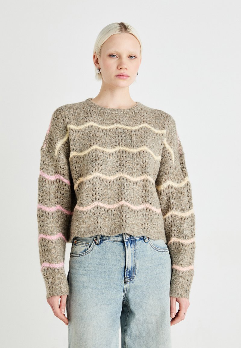 Gebreide trui in lichtbruin met golvende pastelstrepen (roze, geel) en opengewerkte patronen. Ronde halslijn en een cropped zoom. Gecombineerd met lichtblauwe jeans.