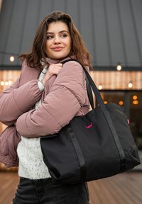 Black tote bag with pink logo and straps, featuring a durable fabric. Model wears a puffy pink jacket and a white sweater, set outdoors.