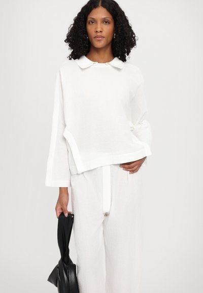 Weißes, strukturiertes Loungewear-Set mit einem kragenlosen, übergroßen Oberteil und passenden Hosen. Ein Model hält eine schwarze Tasche vor einem schlichten Hintergrund.