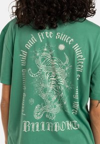 Camiseta de algodón verde con un gráfico blanco de un tigre entrelazado con flores y una serpiente, junto con el texto "BILLABONG."