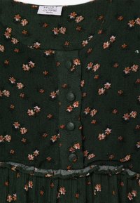 Tissu vert foncé avec un petit imprimé floral blanc et orange, boutons recouverts de tissu disposés en une rangée verticale, et un léger volant en dessous des boutons.