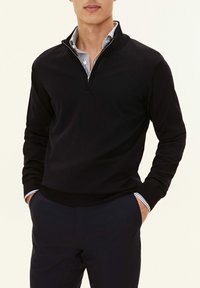 Svart zip-up tröja med långa ärmar och ribbad textur, kombinerad med en randig skjorta med krage under.