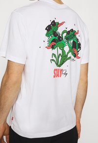 Valkoinen puuvillapaita, jossa on värikäs graafinen kuva vihreistä ja vaaleanpunaisista kasveista selässä, punaisella kirjoituksella "STAY Fly". Lyhyet hihat, rento mitoitus.