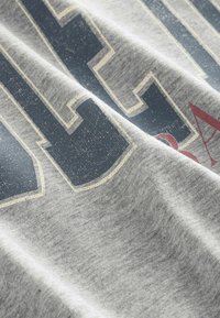 Szara tkanina o miękkiej teksturze z dużymi, zniszczonymi granatowymi literami obrysowanymi w kremowym kolorze oraz małym, częściowo widocznym czerwonym akcentem.