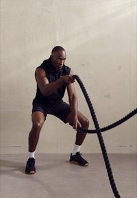 Fekete sportos férfi ujjatlan fekete felsőben és rövidnadrágban vastag harci kötelekkel edz. A háttér világosszürke fal, minimális textúrával.