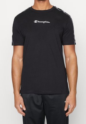 Mand iført sort Champion T-shirt med hvidt logo på brystet og stribedetaljer på ærmerne, kombineret med sorte bukser.