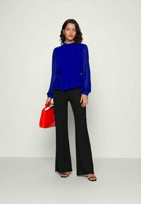 Blusa de mangas longas azul royal com cintura peplum, combinada com calças pretas de corte flare; accessorized com uma mala vibrante vermelha.