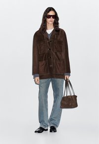 Giacca oversized in suede marrone con quattro tasche frontali, indossata sopra un maglione grigio. Jeans dritti blu chiaro e scarpe nere. Borsa marrone.