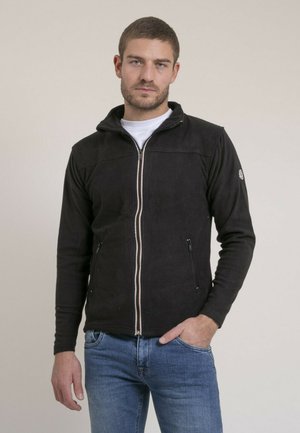 Homme portant une veste polaire noire zippée et un jean bleu, debout avec une main dans la poche sur un fond uni.