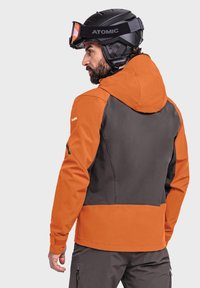Men's ski jacket in orange and dark gray; features a hood, contrasting panels, and a smooth texture. Accompanied by a black skiing helmet.