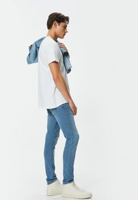 Camiseta de algodón blanca, jeans ajustados de mezclilla azul claro y zapatillas altas blancas. Chaqueta de mezclilla colgada del hombro. Diseño simple, estilo casual.
