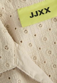 Krämfärgad tyg med blomsterbroderimönster, prydd med en ljusgrön etikett med svart text "JJXX." Mjuk textur; intrikata sömnadsdetaljer.