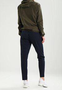 Olivgrön, lättvikts, huvan jacka kombinerad med slim-fit, marinblå, rutig byxor och vita sneakers, visar en avslappnad outfit från baksidan.