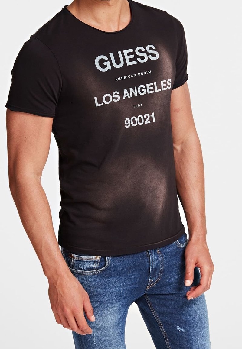 Homme portant un t-shirt noir GUESS avec le texte « LOS ANGELES 90021 », associé à un jean bleu, debout devant un fond uni.