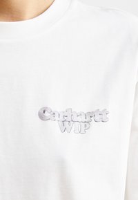 Hvid bomulds T-shirt med et gråt "Carhartt WIP" logo øverst til venstre. Stoflet er blødt med en klassisk runde halsudskæring.