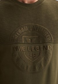 Sudadera verde con material de punto texturizado, cuello redondo acanalado y detalles de logo en relieve. El diseño incluye patrones y acentos.