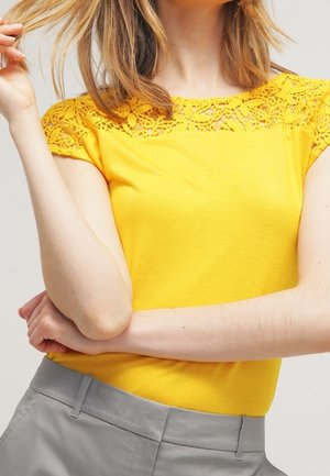 Haut jaune à manches courtes avec des détails en dentelle élaborée au niveau de l'encolure. Tissu lisse et design ajusté associé à un pantalon gris.