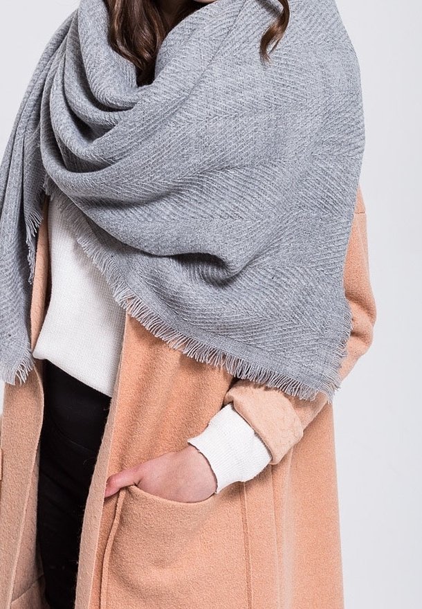 Une personne porte une écharpe texturée gris clair sur un manteau beige avec une poche, un pull blanc en dessous, la main partiellement dans la poche du manteau.