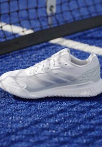 Chaussure de sport blanche avec semelle Cloudfoam+ sur un court de tennis bleu près d’un filet et d’une ligne blanche de délimitation.
