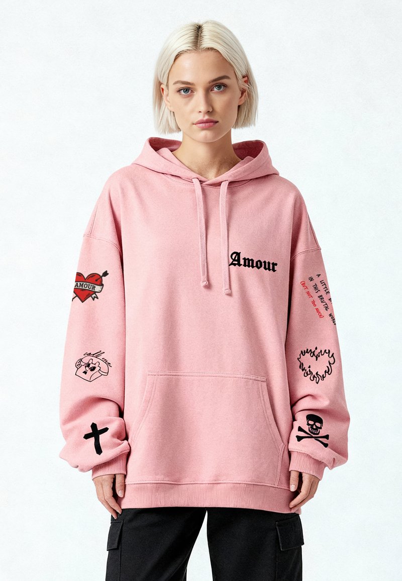 Hoodie surdimensionné rose avec poche avant. Présente des motifs graphiques noirs : texte "Amour", cœurs, un crâne et une illustration de téléphone sur les manches.