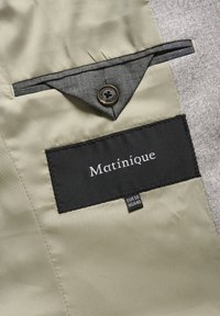 Beige och grått tyg med en svart etikett som säger "Matinique" och en brun knappdetalj på en ficka. Skötsel-taggar synliga.