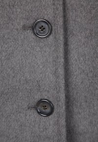 Harmaa villakangas, jossa on kaksi mustaa, neljän reiän nappia, ompelella koristeltu ja pehmeä pinta.