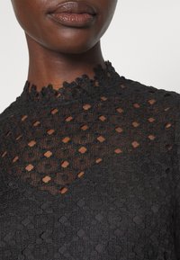 Svart spetstopp med hög halsringning, som har intrikata blommönster och cirkulära mönster, genomskinlig textur och en vågig kant.